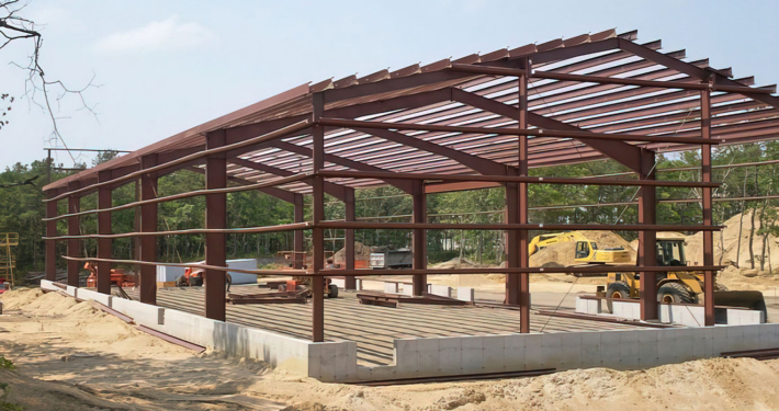 Steel frame of a building under construction on a sandy site, with a bulldozer in the background on a clear day.