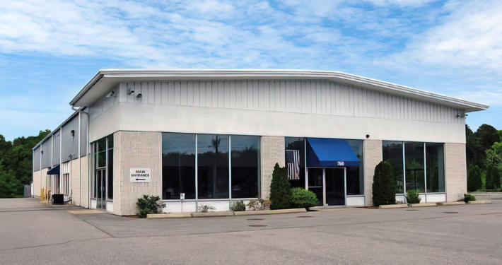 A large, single-story commercial building with white walls, large, dark windows, and a blue awning over the entrance. It is surrounded by a paved parking area and a few shrubs. The sky is partly cloudy.