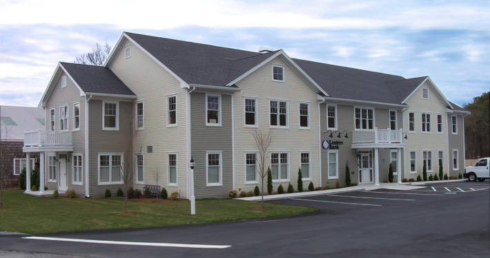 A two-story commercial building with light beige siding and multiple windows. It has a few small trees and shrubs in front, a balcony on the right section, and an adjacent parking lot with a white van parked.
