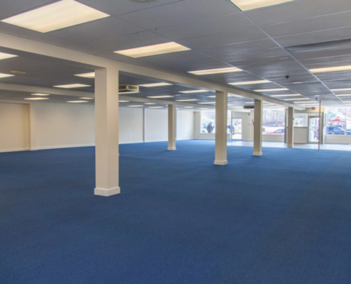 An empty commercial space with blue carpet and white walls is evenly lit by rectangular ceiling lights. Several large windows line one side of the room, allowing natural light to filter in. The area is spacious with visible support columns.