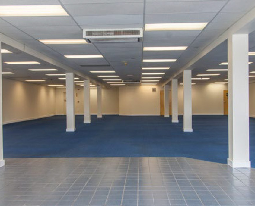 An empty room with blue carpet and white tile flooring near the entrance. The ceiling features fluorescent lights and air conditioning vents. Several white columns are evenly spaced throughout the room, and the walls are painted white.