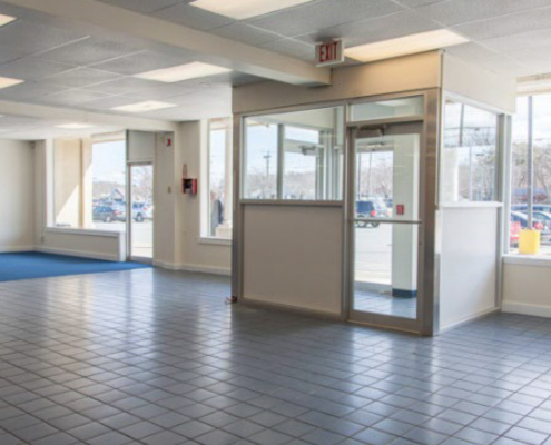 An empty room with large windows, light blue tiled floor, and a small enclosed glass vestibule near the entrance. The room is well-lit by natural light and has fluorescent ceiling lights. An exit sign is visible above the door.