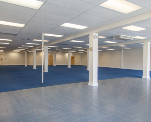 An empty room with white walls, blue carpet flooring, and a tiled entrance. The ceiling has fluorescent lighting, and several white columns are evenly spaced throughout the space.