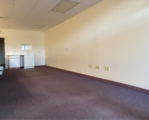 An empty room with beige walls and a brown carpeted floor. There is a small sink and cabinet unit on the left side, under a wall-mounted mirror. The ceiling has recessed lighting panels.