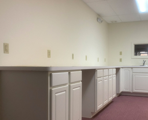 A small room features white cabinets with a long countertop and multiple electrical outlets. There's a sink in the corner, and the room has a carpeted floor and off-white walls. A small window is visible near the ceiling.
