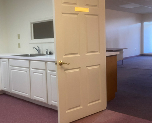 A small room with white cabinets and a sink is visible. The door is partially open, leading to another space with carpeted flooring and a countertop. The walls are light-colored, and there is a small mirror above the sink.