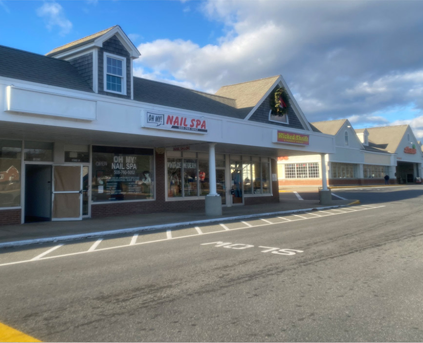 A view of a shopping plaza with a nail spa on the left and a store with a Christmas wreath above the entrance on the right. The sky is partly cloudy, and the parking lot has visible white lines.