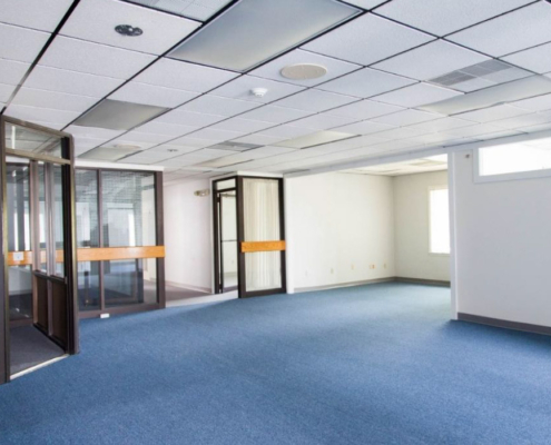 An empty office space with blue carpet, white walls, and a drop ceiling with fluorescent lights. The room has glass-paneled partitions and multiple doorways leading to adjacent rooms. Natural light enters through a window on the right.