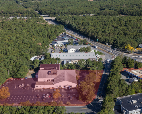 Aerial view of a forested area with roads dividing the land. Several buildings including a large warehouse and other structures are visible.