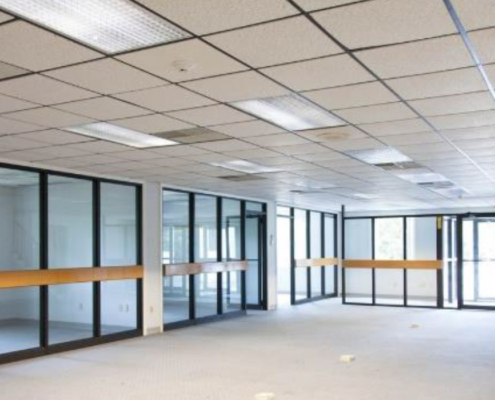 An empty office space with a white grid ceiling and fluorescent lights. The room has glass walls with black frames and wooden paneling. The floor is carpeted in light gray, and there are multiple doorways.