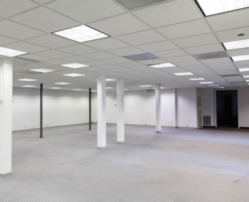 An empty room with white walls and ceiling tiles, featuring several supporting columns. The floor is carpeted in gray, and fluorescent lights are in the ceiling. A darkened doorway is visible in the background.