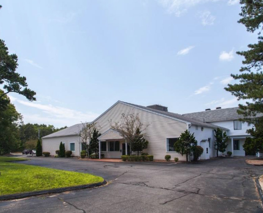 A one-story white building with a sloped roof, surrounded by bushes and trees. It has a paved driveway in front and is set against a clear blue sky.