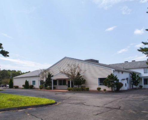 A one-story white building with clapboard and a sloped roof, surrounded by bushes and trees. It has a paved driveway in front and is set against a clear blue sky.