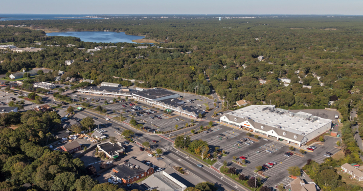 Aerial view of a commercial area surrounded by dense forest. The image shows a large parking lot with several cars, adjacent to a shopping center. In the background, there is a lake, the ocean, and an expansive stretch of greenery.